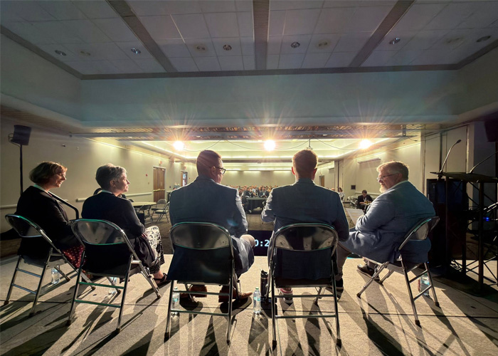 A rear view of NDIT leaders on stage before an event center crowd, their silhouettes enhanced by production lights overhead.