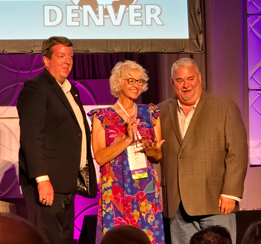 Julie Arp beams while holding her NASTD award while two male colleagues stand with her in support from either side.