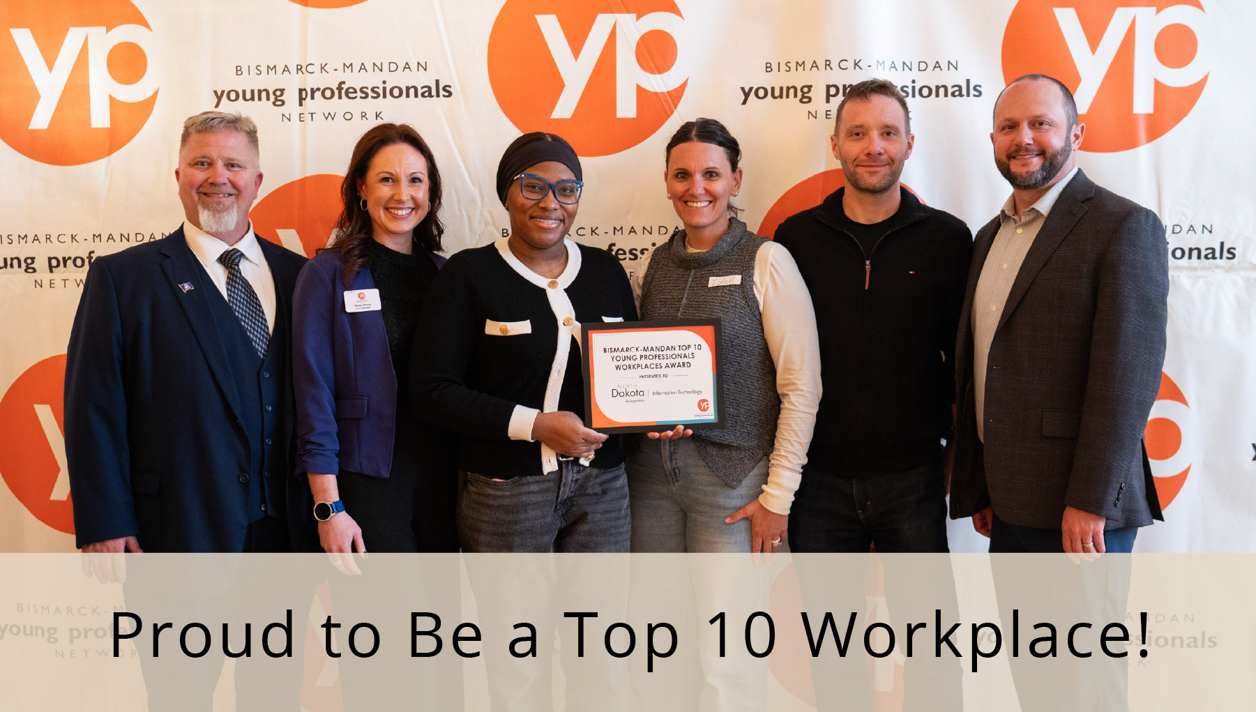 Six excited NDIT team members gather around a plaque, with overlaying text that says we're proud to be a top 10 workplace.
