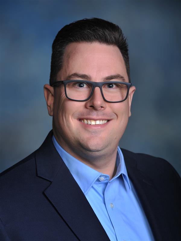A professional headshot of Chris Gergen wearing a dark-blue suit and light-blue dress shirt in front of a charcoal backdrop.