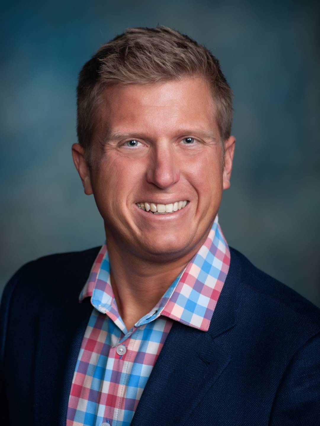 A professional headshot of Corey Mock wearing a dark-blue suit, red, white, and blue, plaid dress shirt in front of a charcoal backdrop.