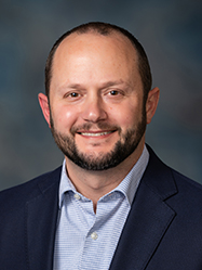 A professional headshot of Craig Felchle wearing a dark-blue suit and light-blue dress shirt in front of a charcoal backdrop.