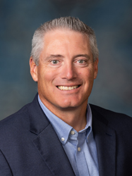 A professional headshot of Greg Hoffman wearing a dark-blue suit and light-blue dress shirt in front of a charcoal backdrop.