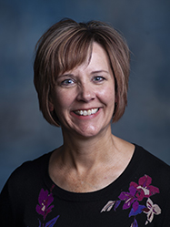 A professional headshot of Kim Weis wearing a dark top with purple flowers in front of a charcoal backdrop.