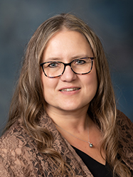 A professional headshot of Shelly Miller wearing a black shirt and outer brown layer in front of a charcoal backdrop.