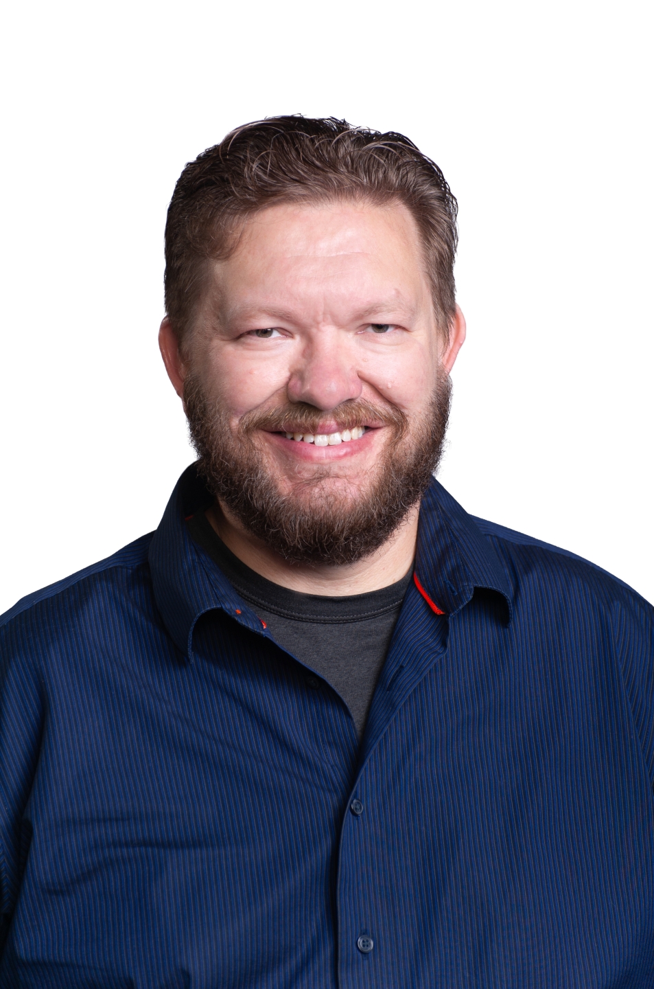 A middle-aged Caucasian man with light brown hair and a beard, wearing a gray t-shirt layered under a dark blue pinstriped button-up shirt.