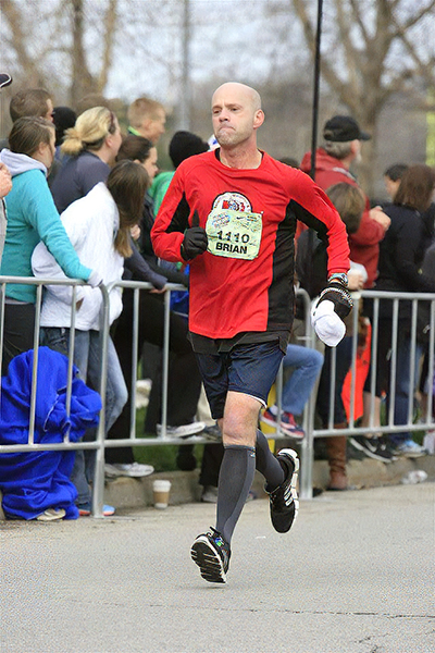 Brian Newby wears a red long-sleeved shirt while participating in a long-distance run with a crowd in the background.