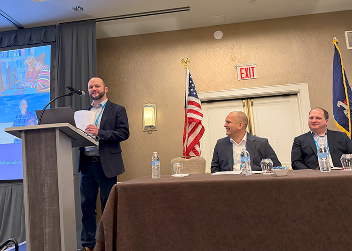 Craig Felchle presents from a podium at a conference while two male colleagues sit at a table to his side. 