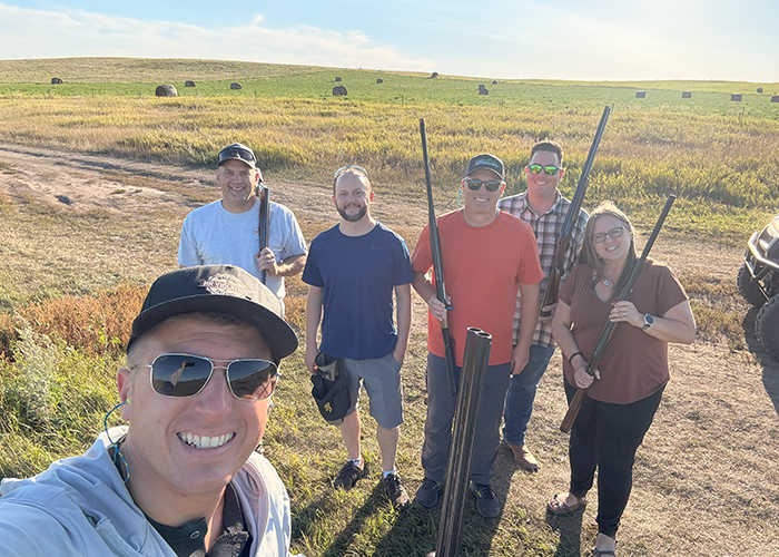 Craig Felchle and other NDIT colleagues gather while holding shotguns at a sporting clay shooting range on a warm, sunny day.