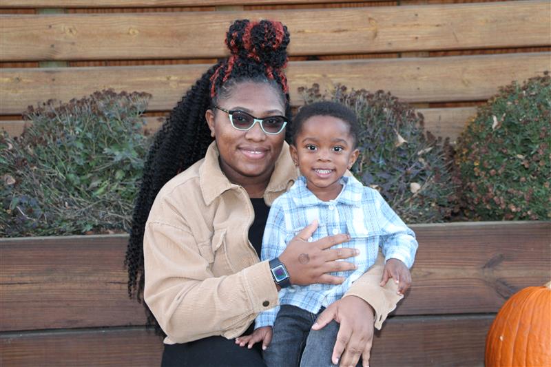 Ingrid Clarke and her young son sit happily together next to a pumpkin on a wood bench with green shrubbery in the background