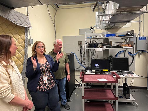 Jessy Wetsch stands between a man and woman in a small technical room while guiding a tour group at the state penitentiary.