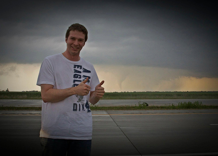 Josh Kadrmas smiles and gives two thumbs up outdoors with a tornado far in the background.