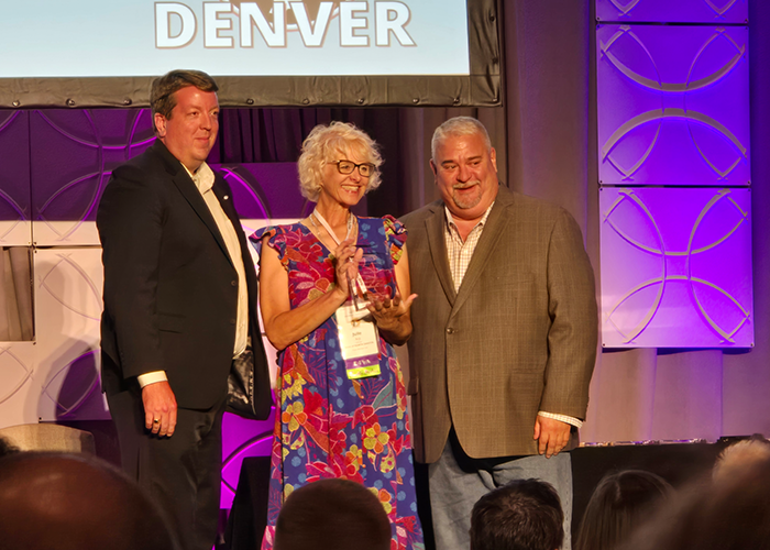 Julie Arp beams while holding her NASTD award while two male colleagues stand with her in support from either side.