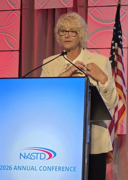 Julie Arp stands behind a podium and does heart hands over her heart while speaking at the 2026 NASTD Annual Conference.