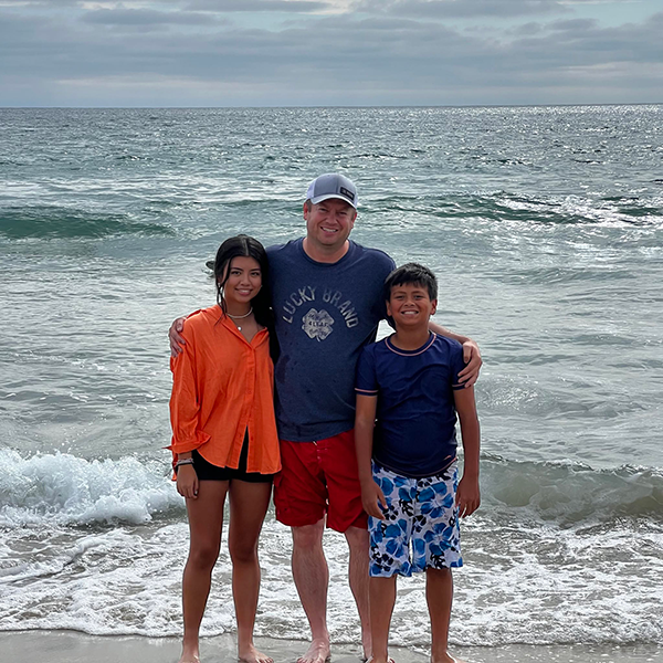Mike Benz smiles and poses in front of an ocean with his young son and daughter on a calm overcast day.