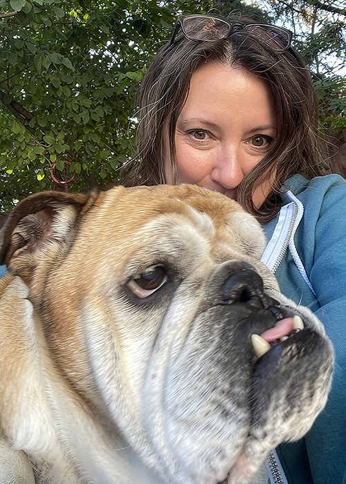 Monique Durgin, wearing a light-blue hoodie and reading glasses on her head, takes an outdoor selfie with her English bulldog, Winston.