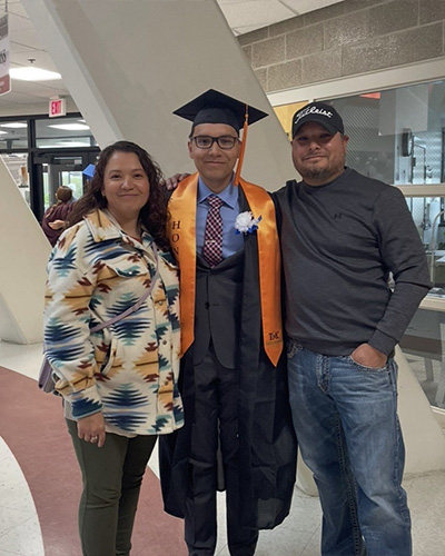 Quentin Brien, wearing a black graduation gown with an orange honor sash, poses for a picture between his parents.