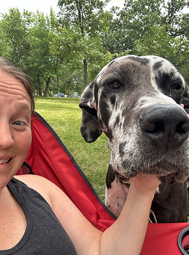 Raulie Berthold sits on a lawn chair and smiles while petting her gray-and-white Great Dane on a beautiful day.