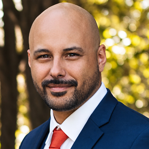 A professional headshot of Jared Lemieux standing in front of a tree while wearing a blue suit, white shirt, and red, orange tie.