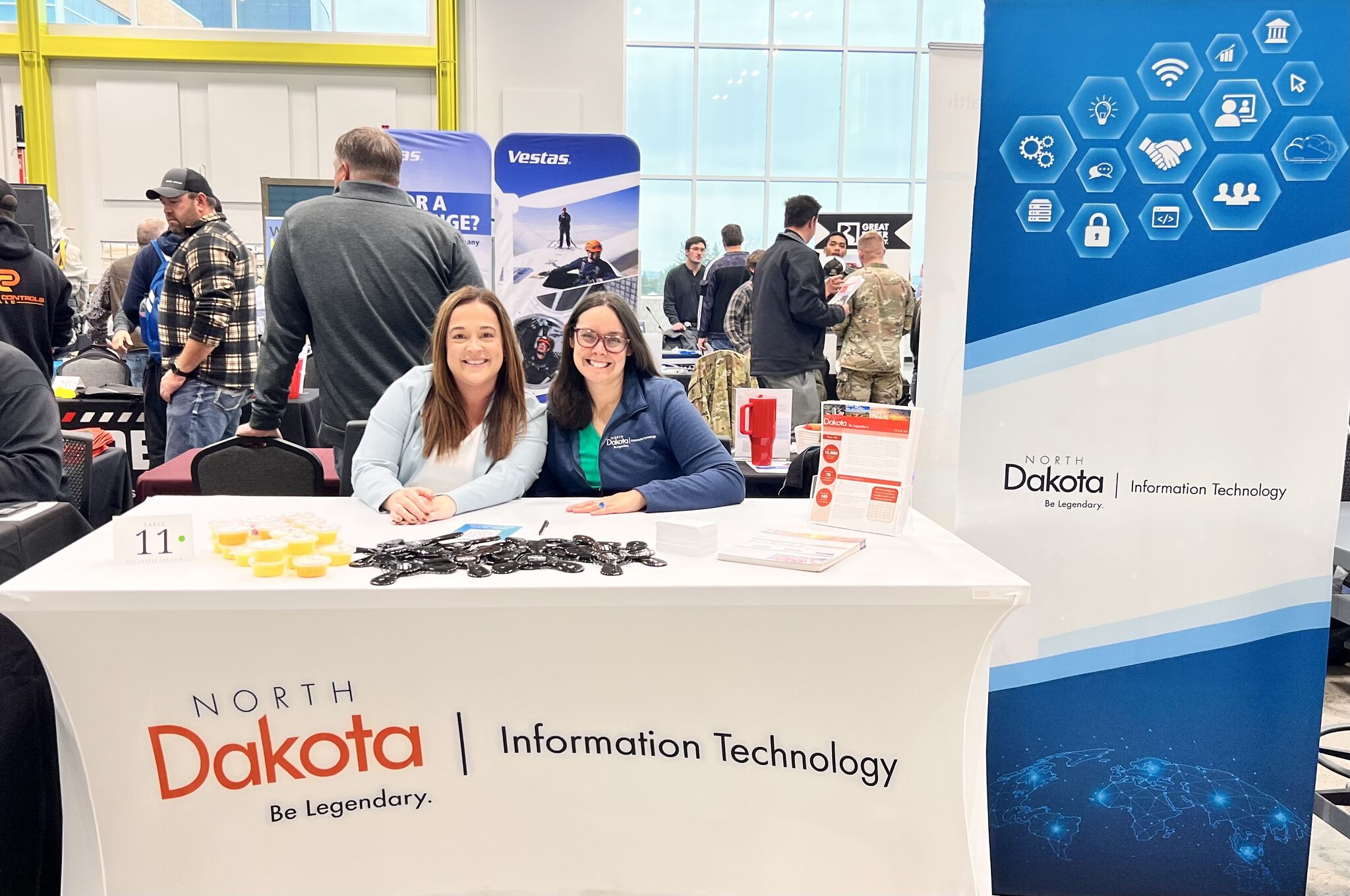 Colleen T. and a female colleague huddle happily together for a picture while sitting behind an NDIT-themed job fair booth.