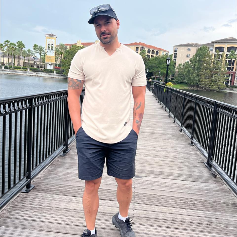 Jared Lemieux wearing a t-shirt, shorts, and hat with his hands in pockets walking across a pedestrian bridge on a sunny day.