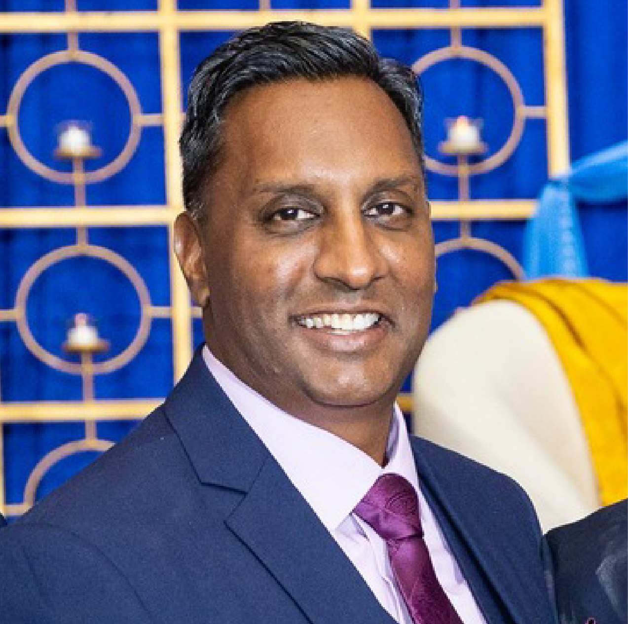 Professional headshot of Ravi Shivdat smiling while wearing a blue suit, light purple dress shirt, and dark purple tie.