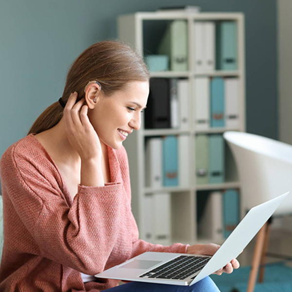 A happy young woman wearing a hearing aid adjusts her hair while sitting down on a couch and using her laptop.