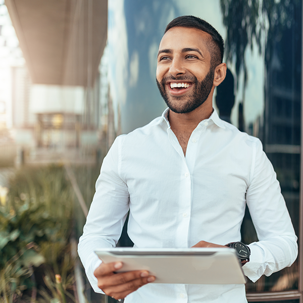 A happy man wearing a white dress shirt works on his tablet while standing in front of glass windows outside.