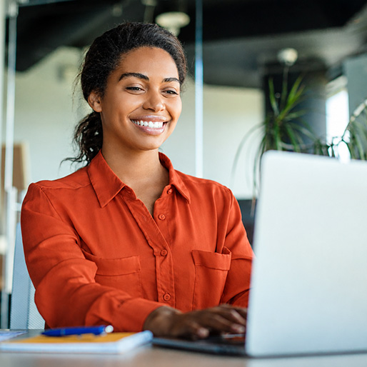 Happy woman wearing and orange blouse sitting at a desk with a laptop typing.
