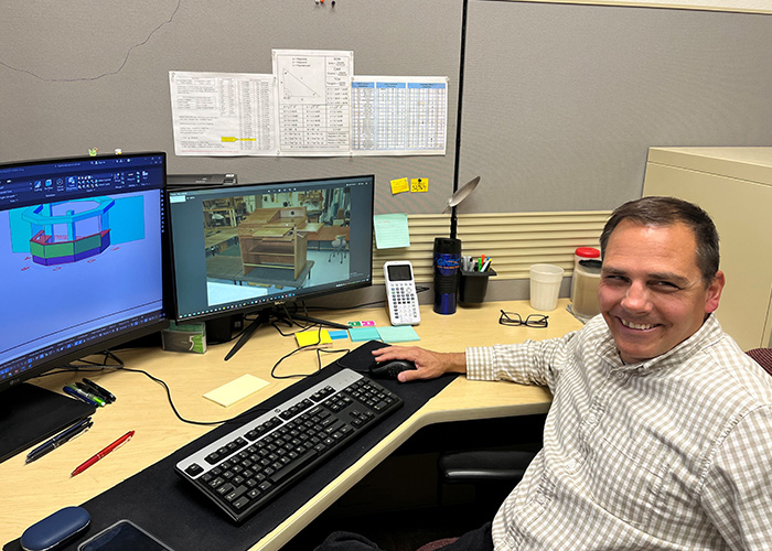 Dave Sission smiles while sitting at his work desk, with two monitors featuring the AutoCAD system he works in every day.