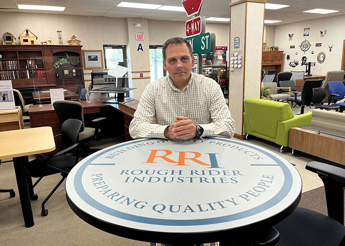 Dave Sisson sits at a round table featuring the Rough Rider Industries logo, surrounded by furniture on display.
