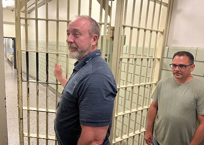 A smiling man (Rick Gardner) opens a prison door while another man (Dave Sisson) stands off to the side in a well-lit hallway.