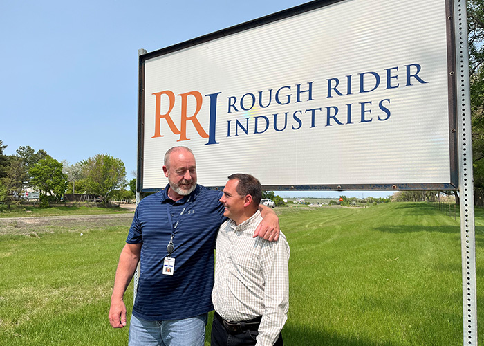 Rick Gardner and Dave Sisson smile while standing arm in arm in front of the Rough Rider Industries sign in Bismarck, ND.