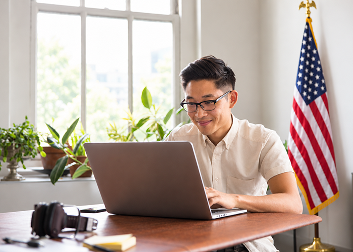 Male with a white collared shirt and glasses sitting at a desk with a laptop smiling.