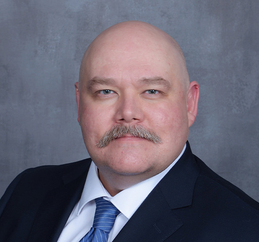 A professional headshot of John Rodenbiker, Director of Cyber Operations, wearing a dark suit.