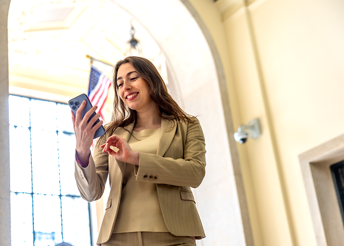 Young woman in a tan suit jacket and tan shirt holding a cell phone smiling.