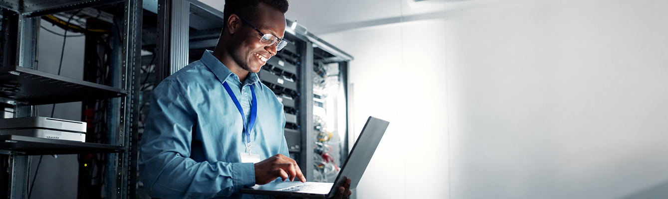 A happy man wearing a light-blue dress shirt and glasses stands in a server room while holding a laptop computer.