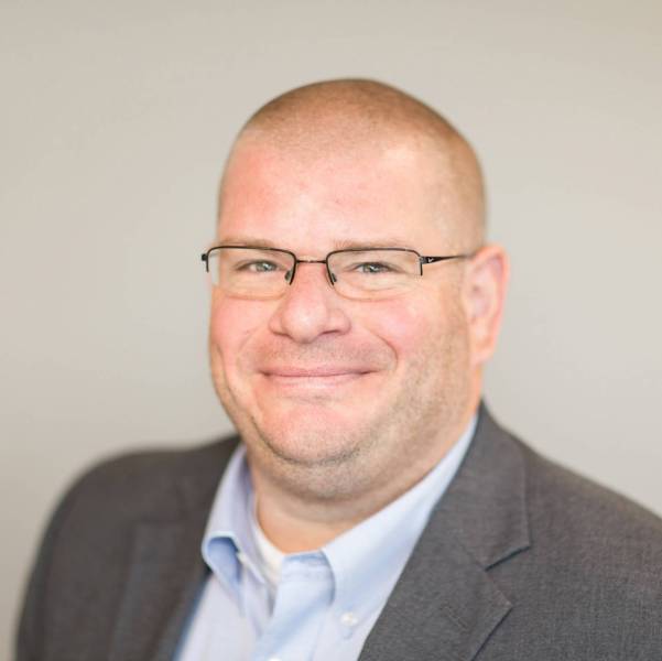 A professional headshot of Andrew Sherwood wearing glasses dressed in a light blue collared shirt and dark grey suit jacket.