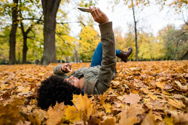 A young woman wearing a green jacket and jeans holds her smartphone in the air to take a selfie while laying down in leaves