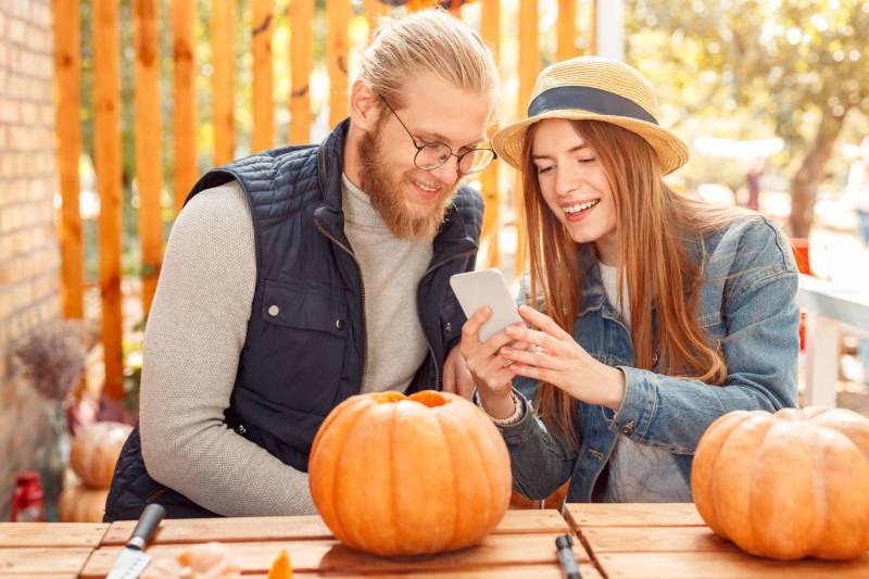 A happy young man and woman look at a smartphone while taking a break from carving pumpkins on a colorful fall day