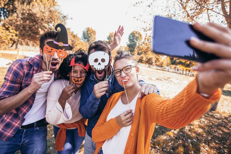 A happy young group of friends hold Halloween masks up to their faces while taking a fun selfie on a bright autumn day