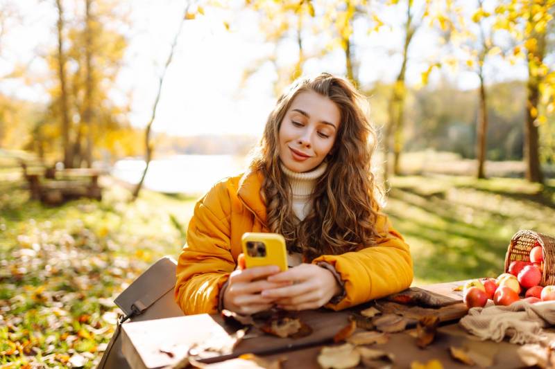 A smiling young woman looks at her phone while sitting at a picnic table next to a basket of apples on a bright autumn day