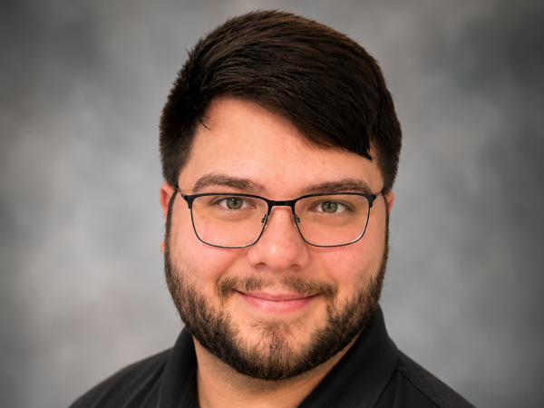 A professional headshot of Skyler Lunsford wearing a black polo shirt and glasses