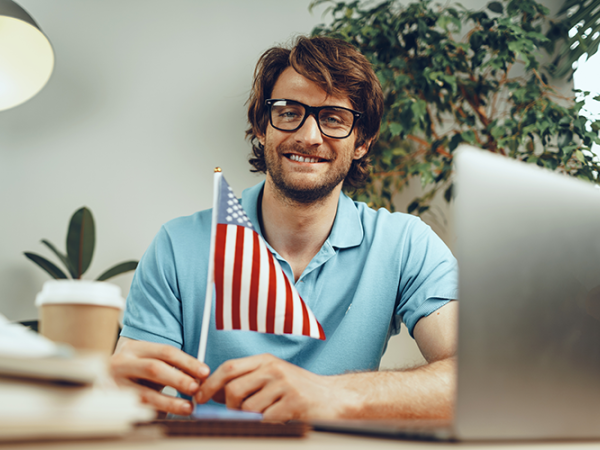 Man with glasses and a blue polo shirt sitting at a desk with an American flag.