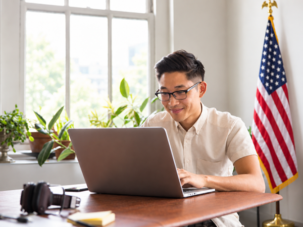 Male with a white collared shirt and glasses sitting at a desk with a laptop smiling.