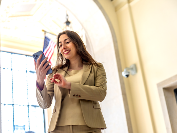 Young woman in a tan suit jacket and tan shirt holding a cell phone smiling.
