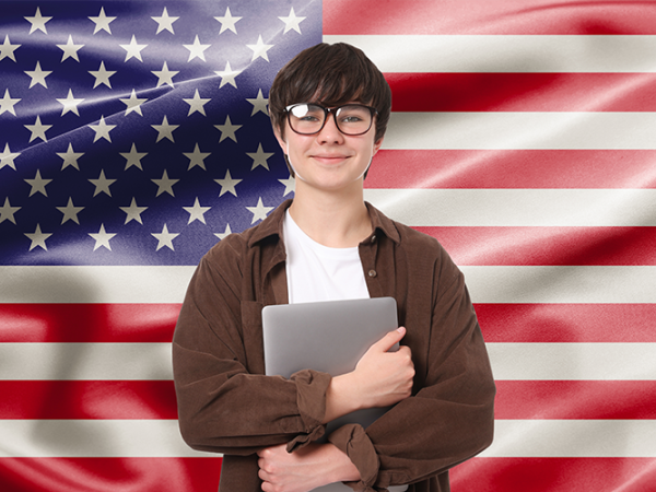 Young male standing in front of the American flag with a laptop.