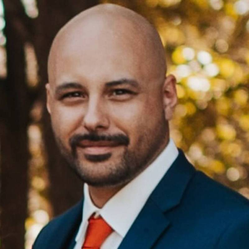 A professional headshot of Jared Lemieux standing in front of a tree while wearing a blue suit, white shirt, and orange tie.