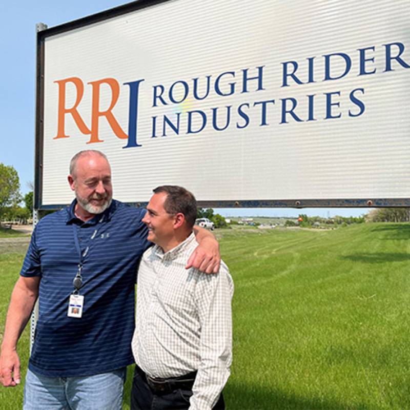 Rick Gardner smiles and puts his arm around Dave Sission in front of the outdoor Rough Rider Industries sign in Bismarck, ND.