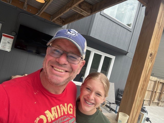 Andrew Sherwood and his daughter smile while taking a selfie together under a wooden outdoor deck attached to a gray building